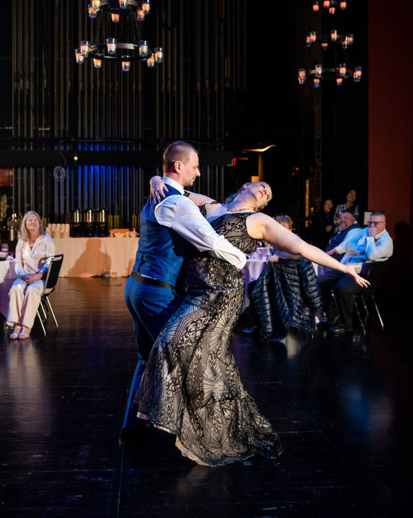 A man in a blue suit and a woman in a beige and black dress with light complexion dancing together.