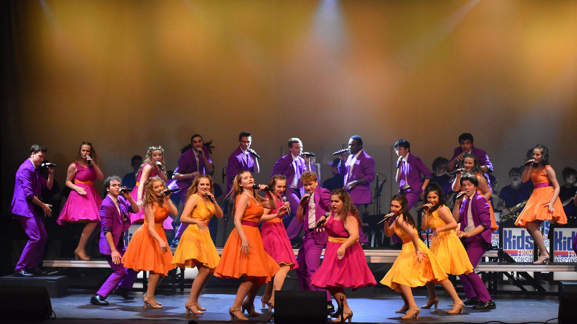 Kids from Wisconsin performing on stage. in two large lines. Some are wearing purple suits and some are wearing red, yellow, or orange dresses.