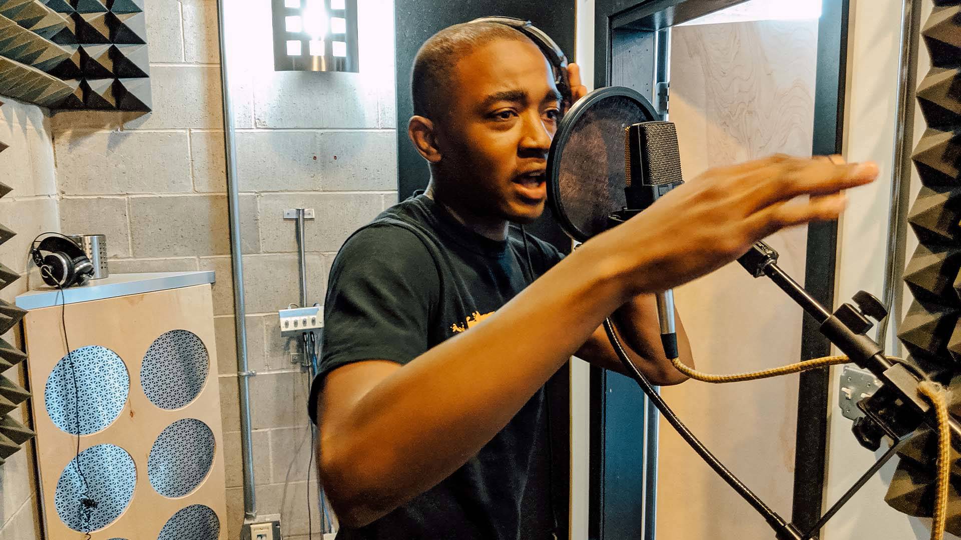 A young Black man standing in a sound booth recording vocals into a microphone.