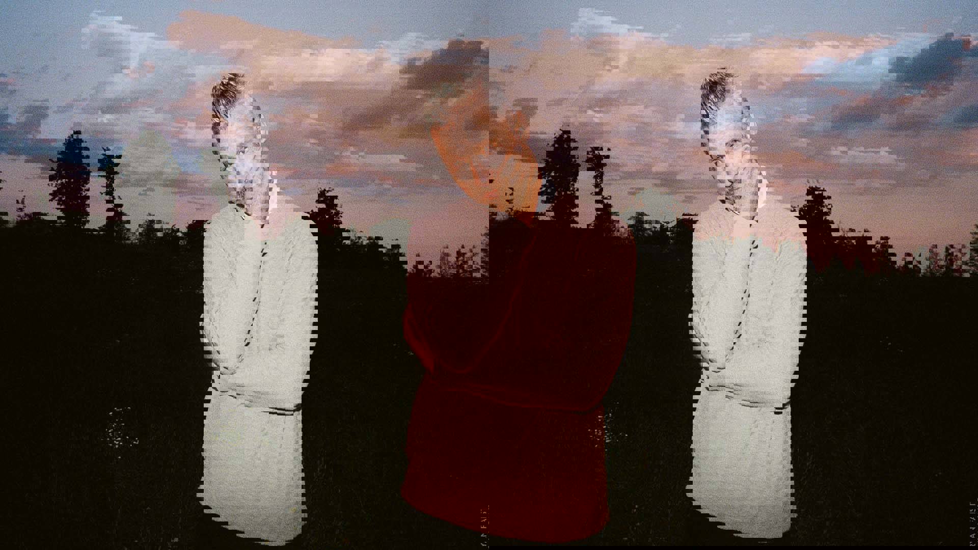 A middle-aged caucasian man with short gray-white hair and trimmed facial hair. He is wearing a dusty pink sweater over a white collared shirt with dark pants with his left arm around his chest and his right hand pressing his left cheek. He is standing outside with a forest and pink sunset behind him.