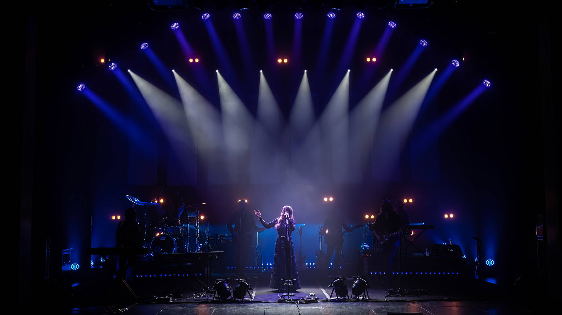 Rumours of Fleetwood Mac band performing on stage. A caucasian woman with long blonde hair stands in the center at a mic stand, singing with multiple spotlights directed at her. She is wearing a dark, flowing long dress and her right hand is lifted in the air.