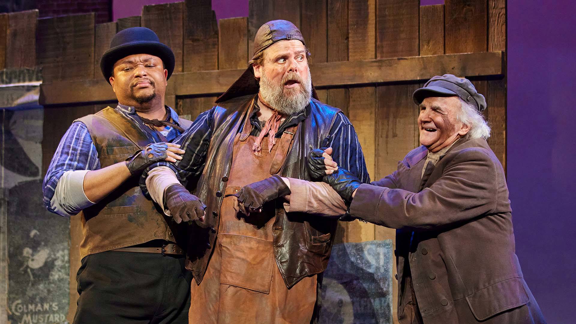 Two men link arms with a third man in front of a wood fence. All three wear brown and dirty clothing. 