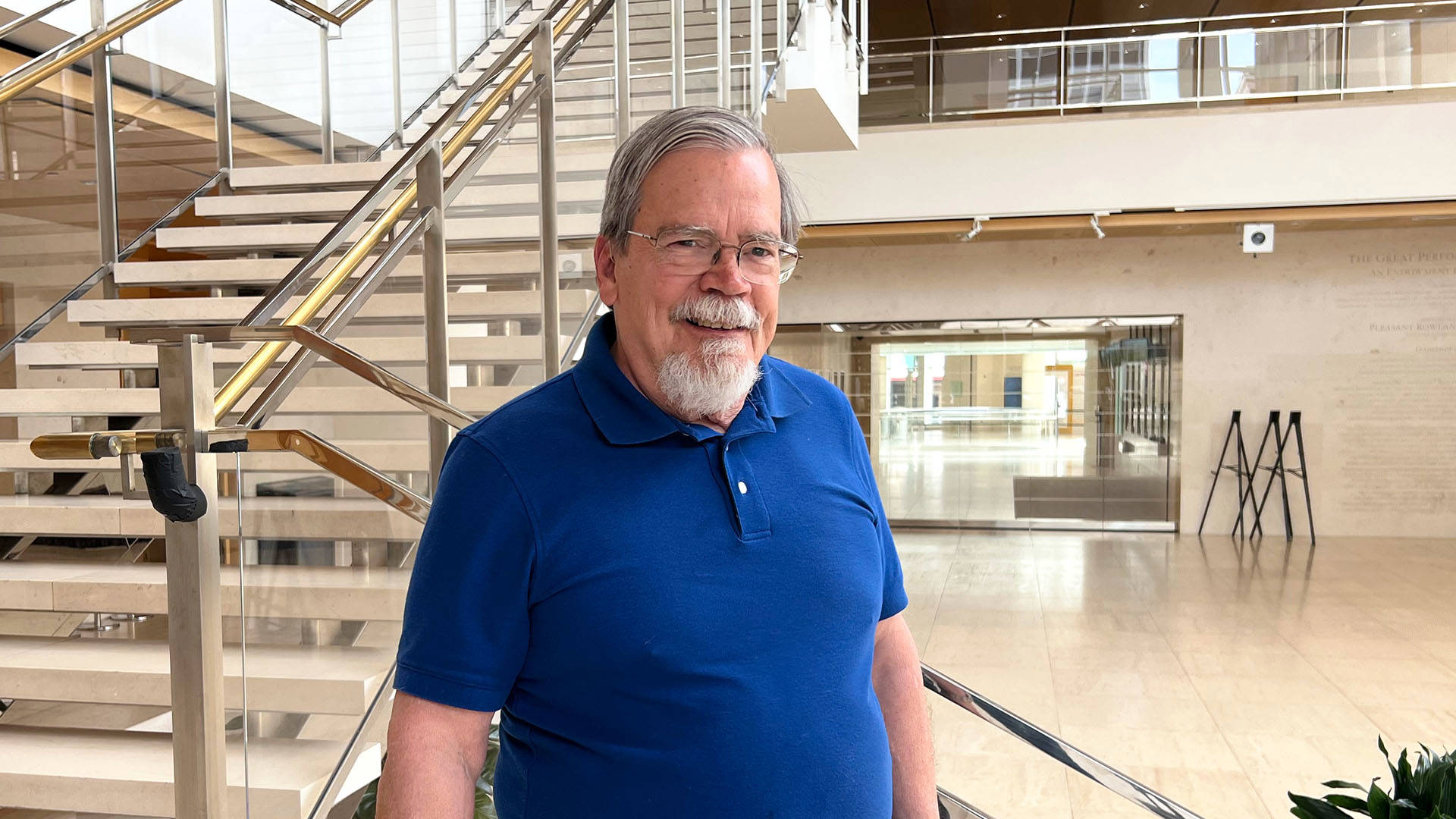 Steve Schroeder, an older white man with a medium-short grey and white hair, a white goatee and silver-framed glasses. He is wearing a blue polo, smiling in Overture Hall main lobby.