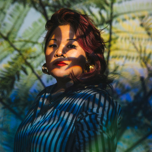 A photo of Leslie Damaso, a young woman with medium complexios, reddish dark hair, and brown eyes is a striped blue and black shirt. Her face is cast in thin shadows.