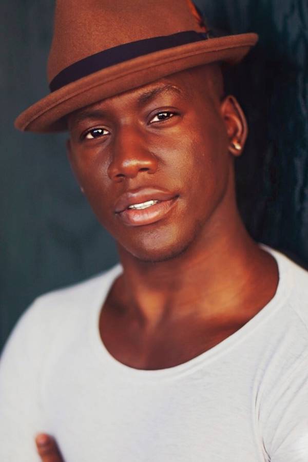 Headshot of a young man with dark complexion, brown eyes, a white shirt and a brown rimmed hat on.