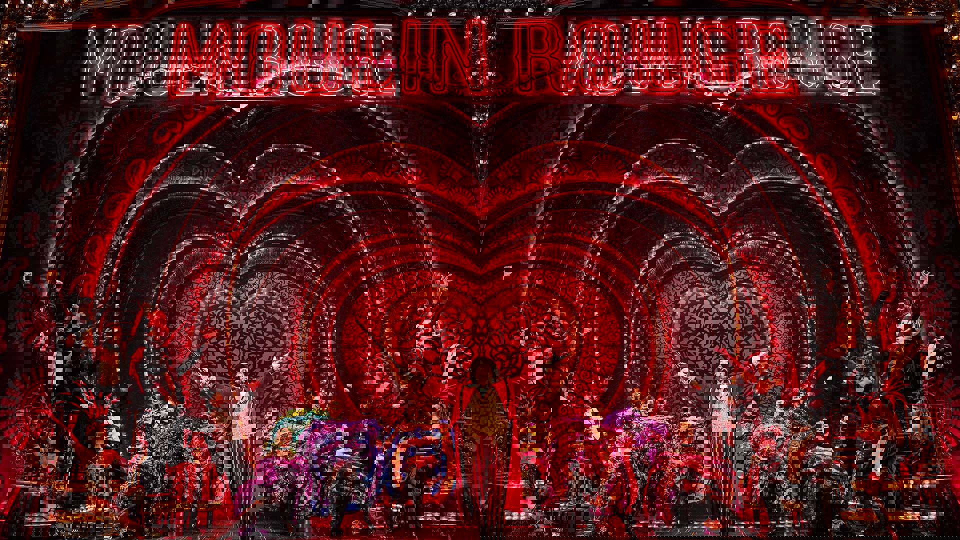 The cast of Moulin Rouge! in various colorful costumes performing together in front of a red heart shaped set.