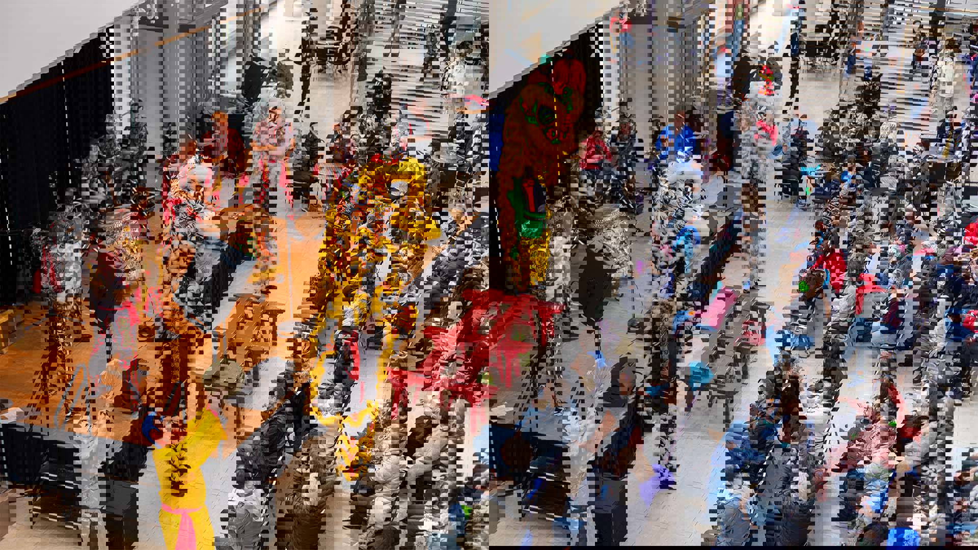 A wood stage set up in Overture Hall main lobby with a seated audience watching a Chinese drumming band perform with pairs of people dressed as Chinese dragons with yellow fur, green, black and red ornate accents and large eyes..