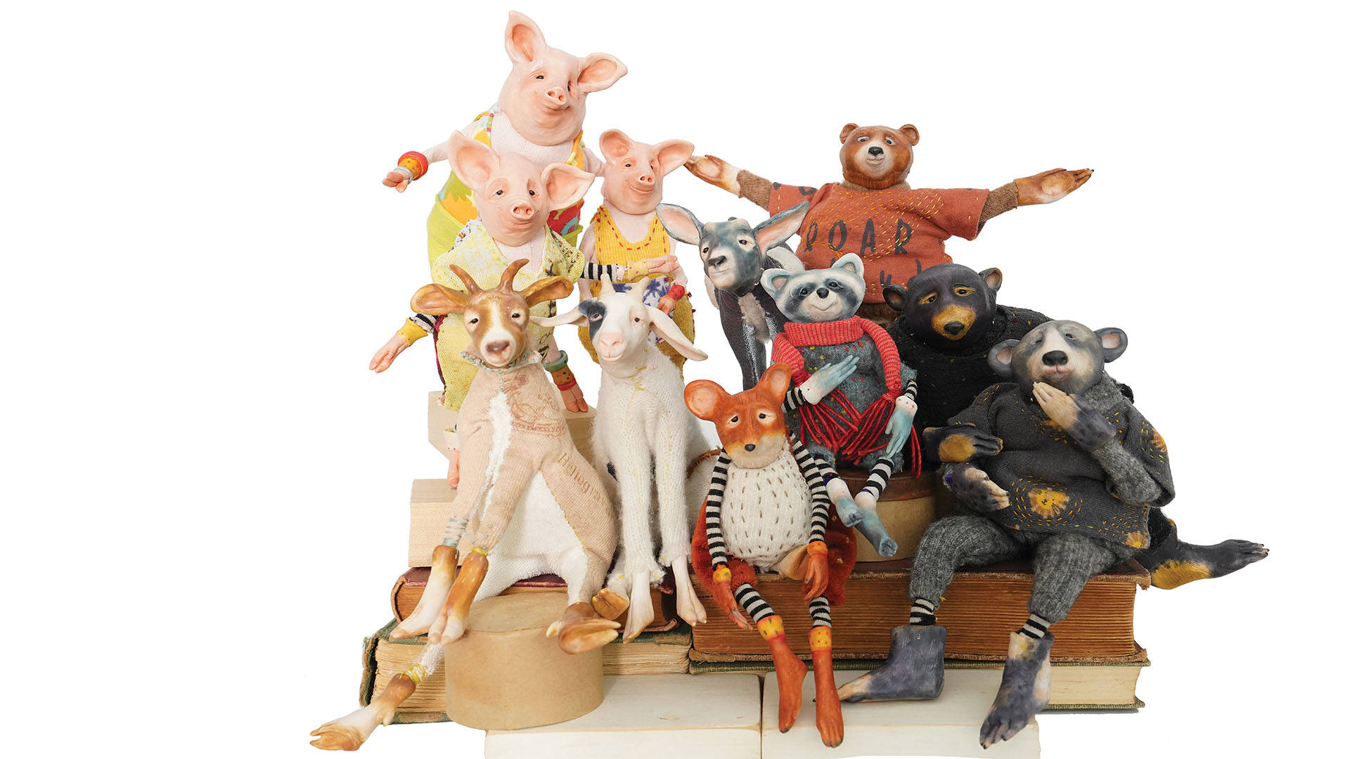 A white banner depicting a display of plastic, realistic animal toys of 3 pigs, 2 goats, a fox, bears , a raccoon and a donkey, wearing knitwear clothing, sit together on a stack of books.