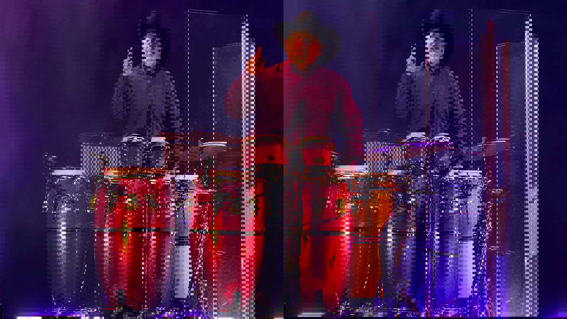 A drummer wearing a black cowboy hat standing with a red set of hand drums.