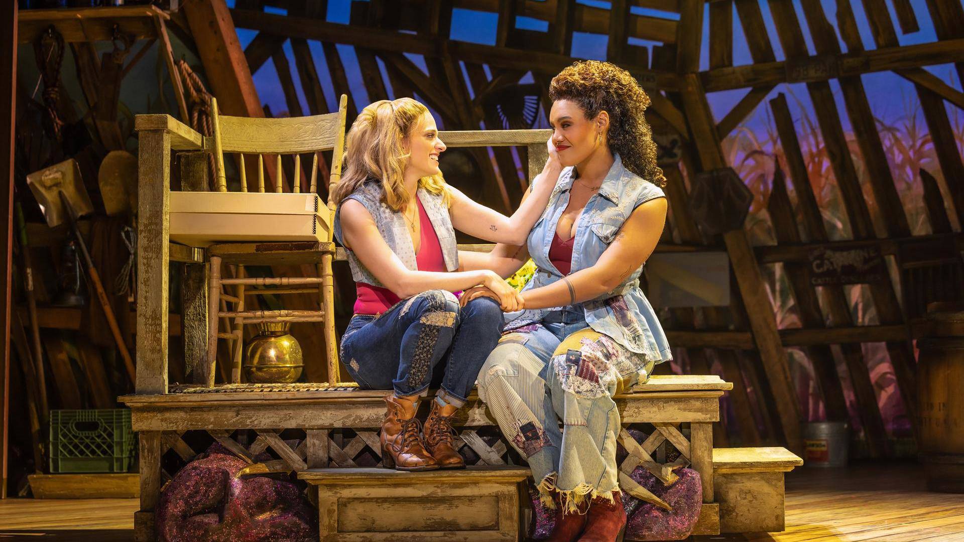 Two woman sitting next to eachother and touching eachother warmly. The woman on the left has blonde hair, light complexion, jeans and a vest over pink top. The woman on the right has medium complexion, curly hair , pants with many patches, and a blue vest.