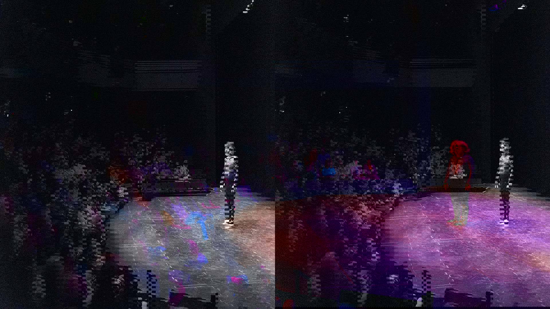 A man with light complexion, a red, long hair wig, and a black jumpsuit stands in front of an audience.