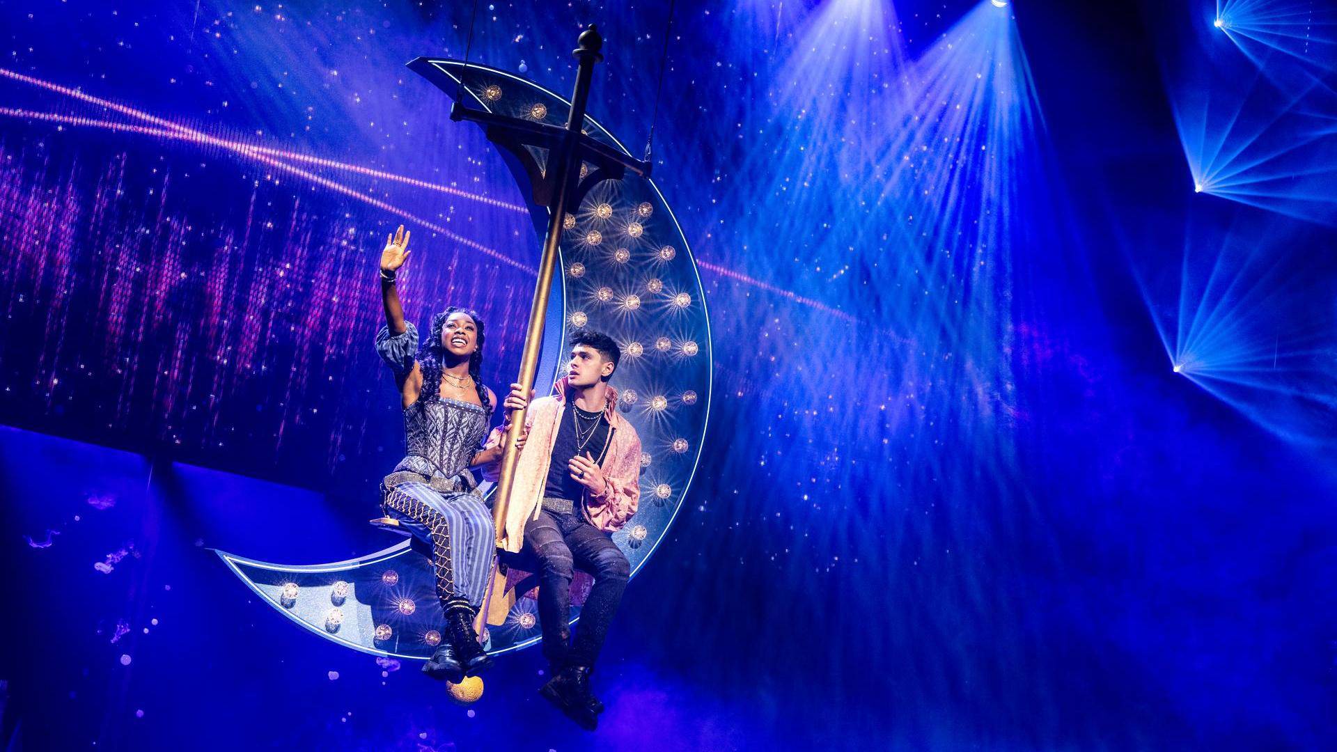 Two people sitting on a moon-shaped set piece suspended in the air. The surround lights are blue and purple.