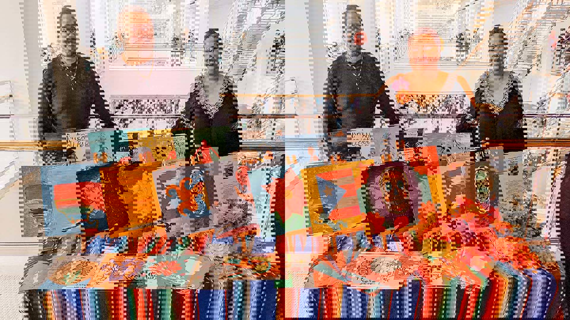 Two people both with medium complexion and dark long sleeve shirts standing behind a long table with a striped colorful tablecloth displaying small paintings at The Latino Art Fair.