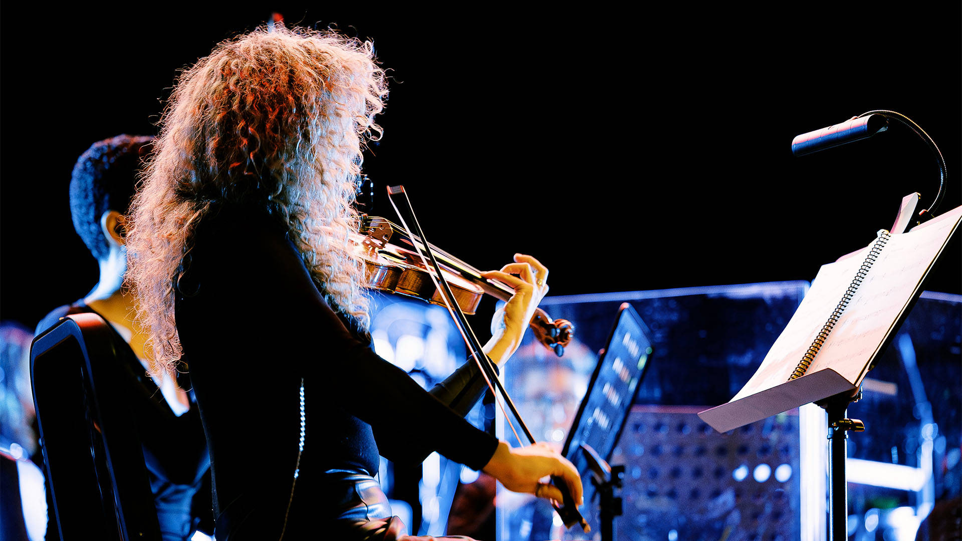 A woman with long curly blond hair and a dark outfit playing violin and looking at a music stand.