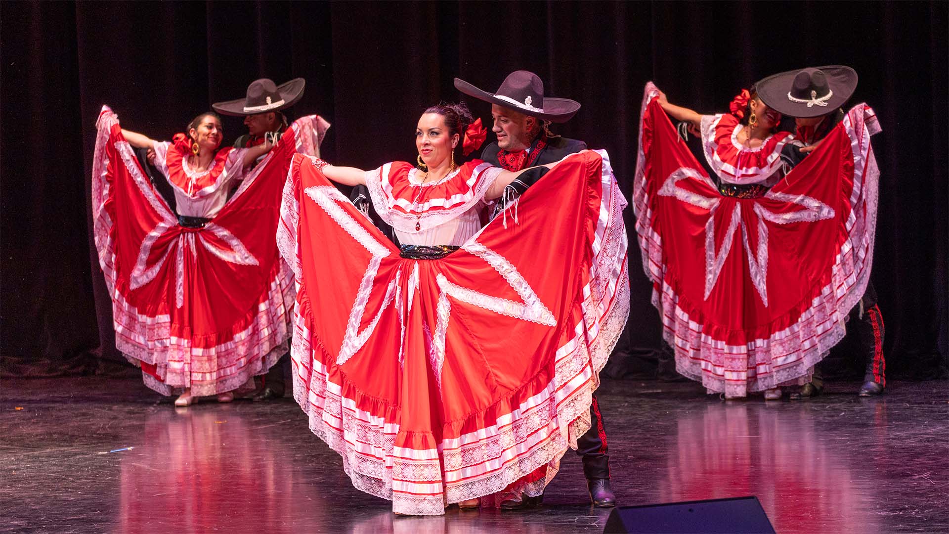 Three women with medium complexion wearing vibrant red dresses holding out their skirts with both arms wide to display the skirt fabric. Each have a male partner in black with black sombreros behind them.