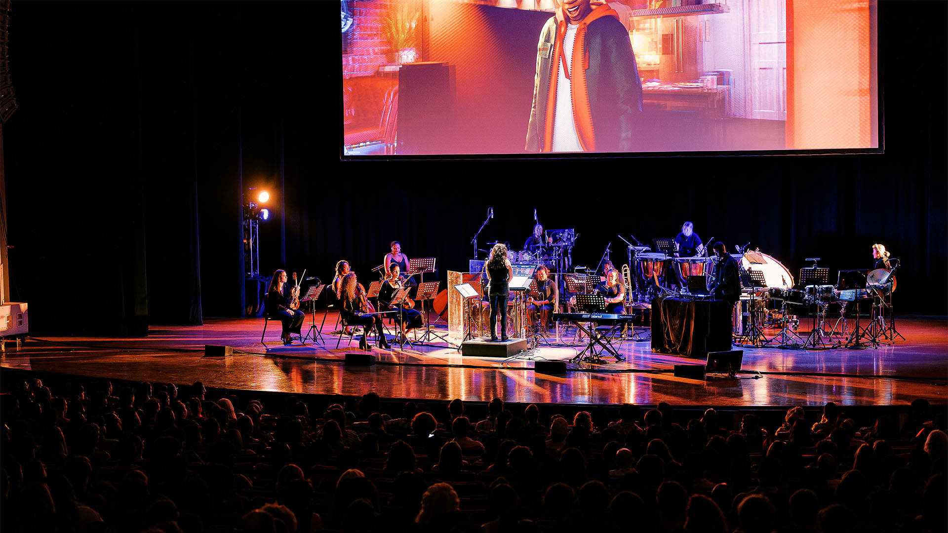 An orchestra on stage performing with Spiderman: Into the Spiderverse playing above on a large screen
