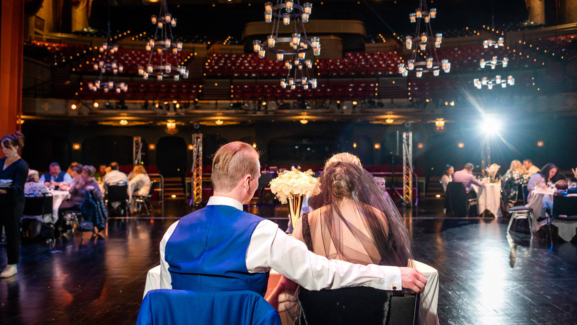 A man in a blue suit and a woman in a black dress sitting facing away from the viewer. They are on Capitol Theater stage and gazing at the rest of a wedding party.