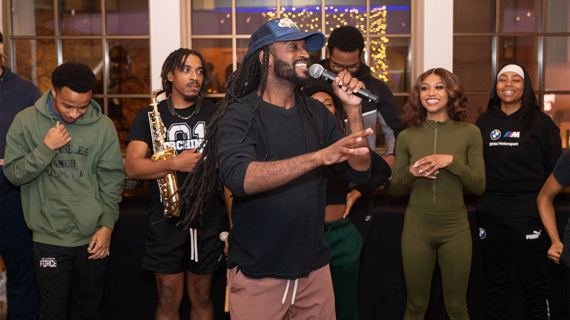 A young african american man with with long dark hair and baseball cap speaking into a microphone in front of six other performers from drumline live