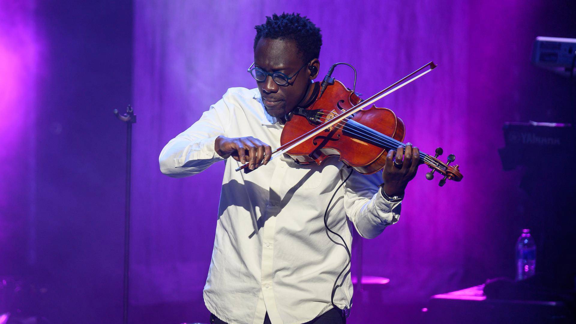 A man with dark complexion, a white shirt glasses concentrates as he plays violin. The background is purple.