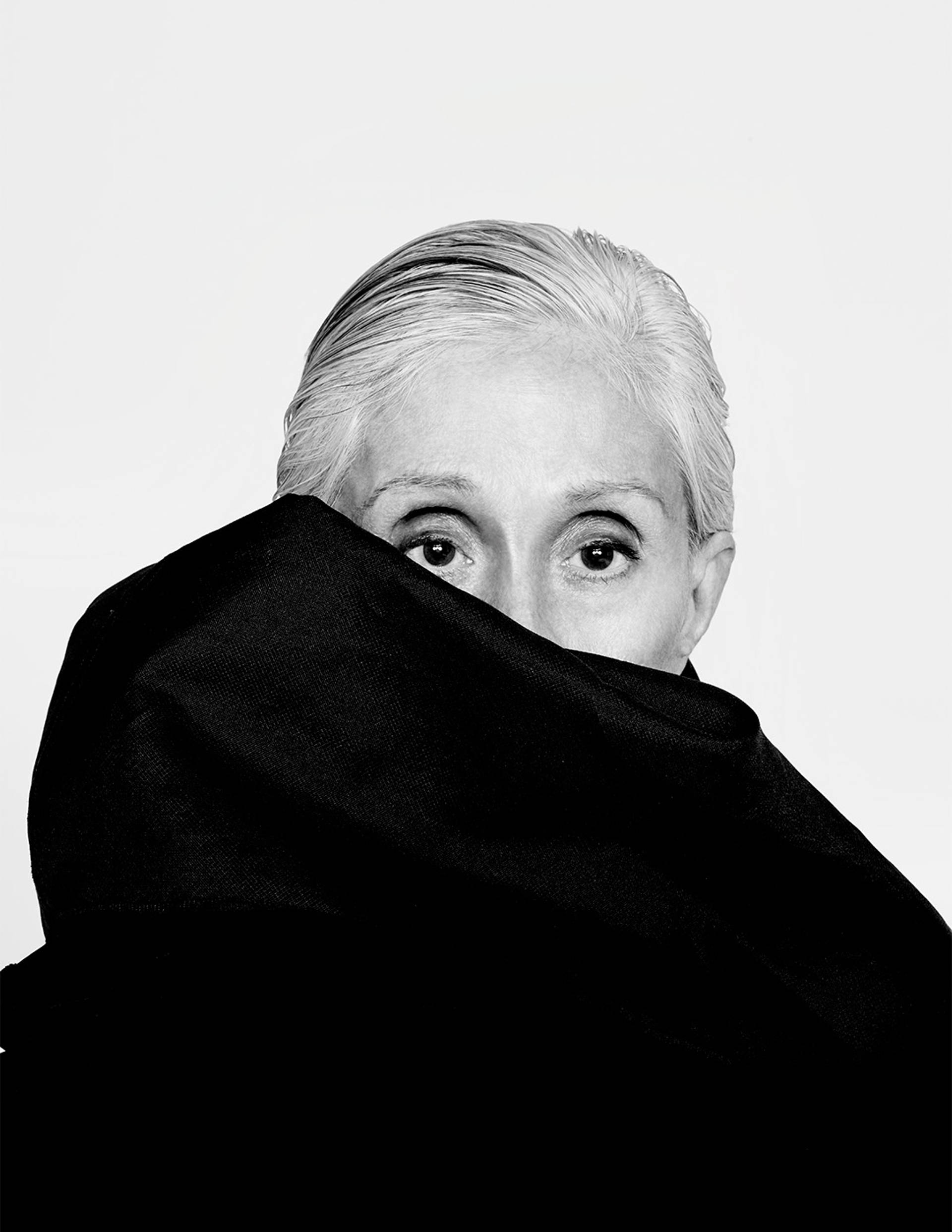 Twyla Tharp headshot in black and white. An older caucasian woman with white hair slicked back and large black scarf that obscures her face from below the eyes.