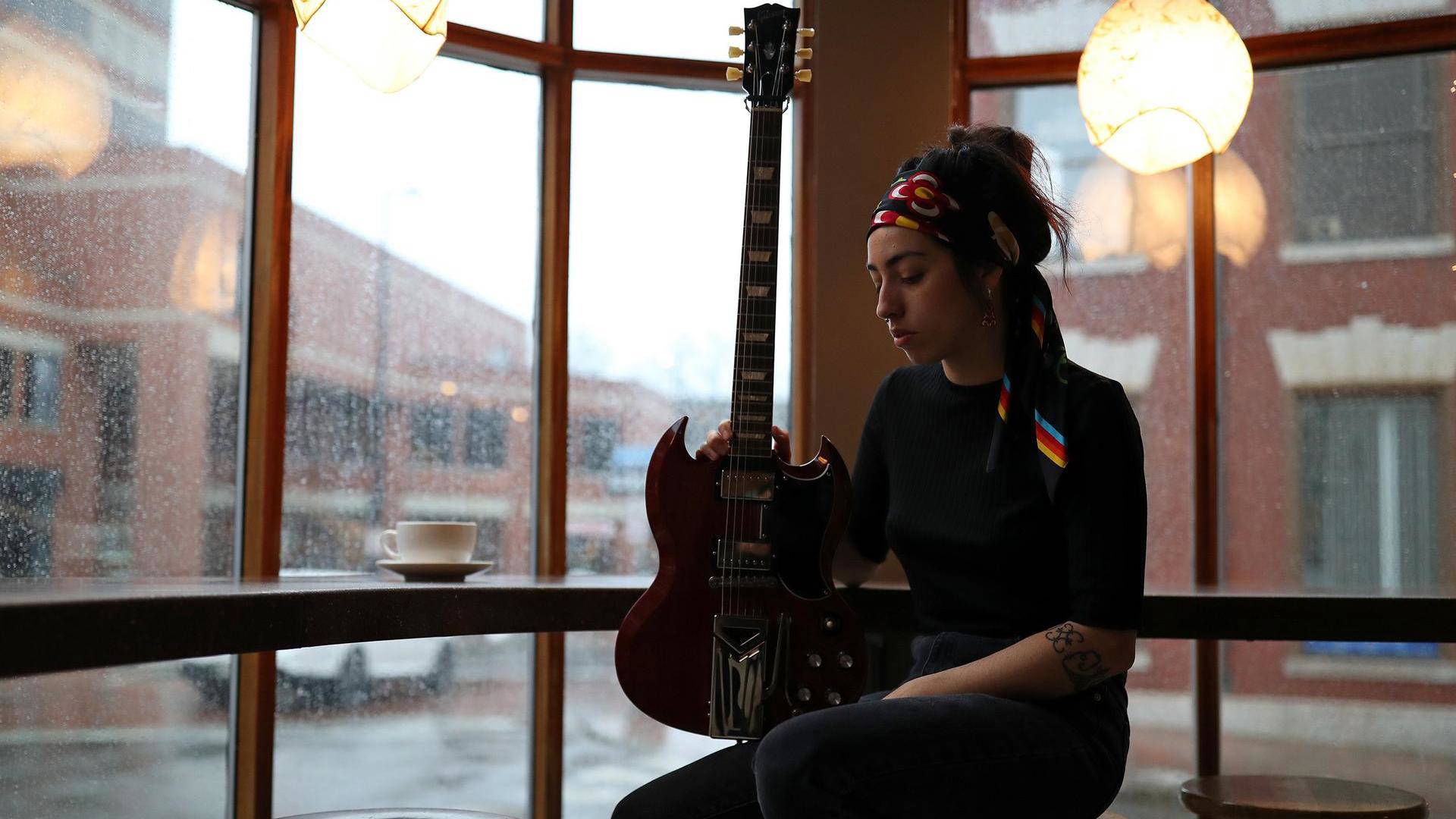 A young woman with medium complexion, long brown hair, and a colorful head tie sitting in front of a window with a red electric guitar resting on her knee.