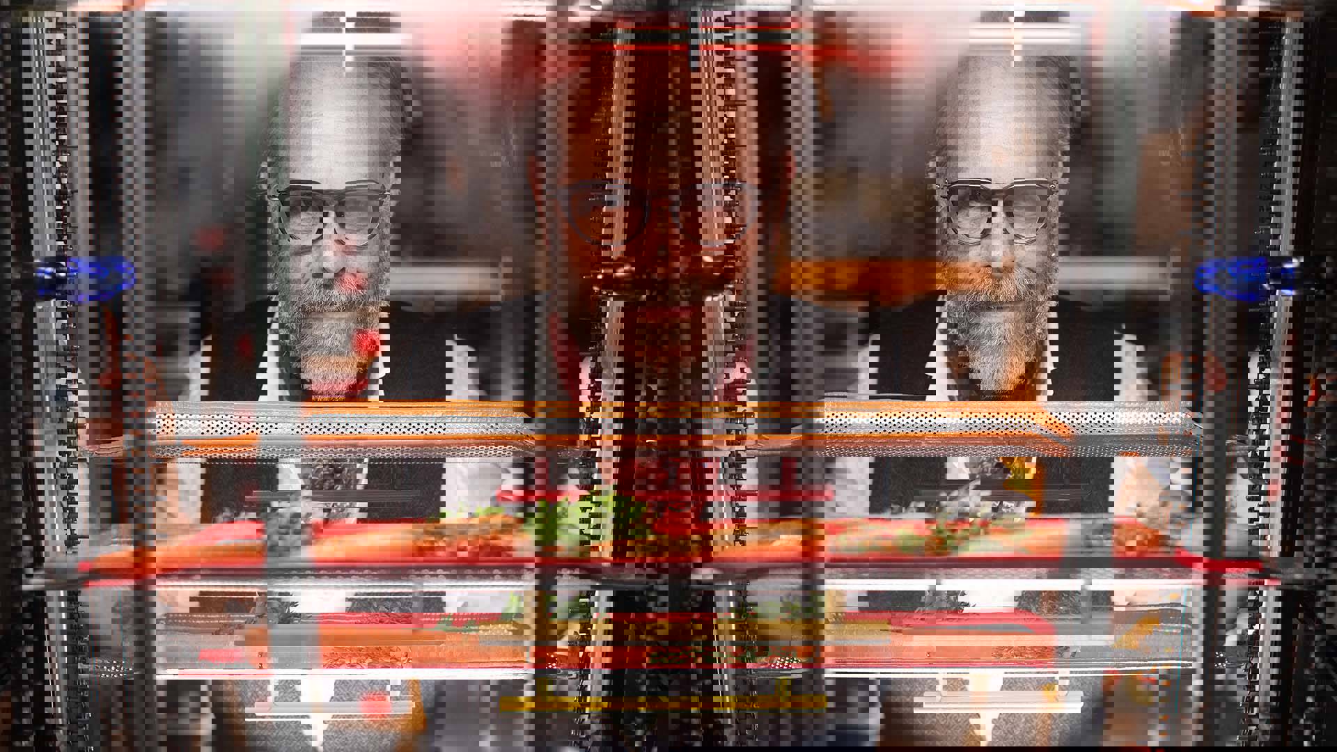 Alton Brown, a middle-aged caucasian man with a gray-brown beard and receding gray-brown hair in black framed glasses, standing behind a clear glass panel with shelves containing orange carrots. He has a smirk and is wearing a white collared shirt, black vest and red handkerchief cravat. 