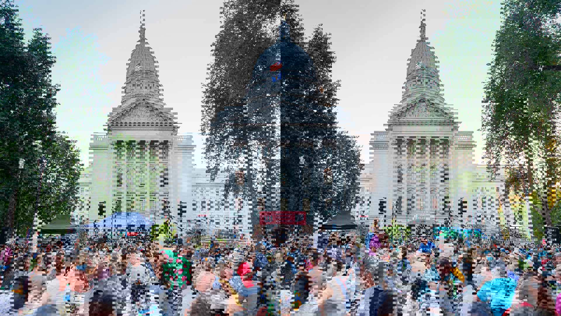 The Wisconsin state capitol as the sun is setting. A stage is set up with an orchestra. Hundreds of people sit outside socializing and listening to the music.