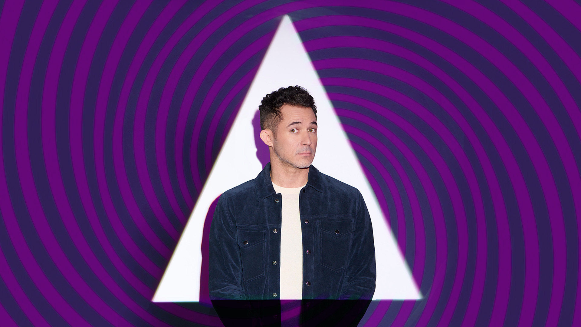 Justin Willman, a caucasian man with short brown hair and brown eyes wearing a white shirt and an open blue button up shirt. He is enclosed in a white triangle and the background is a spiral of purple and blue colors.