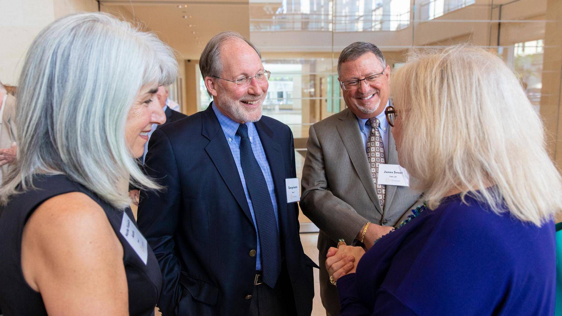 Four people in business attire stand smiling and talking to eachother.