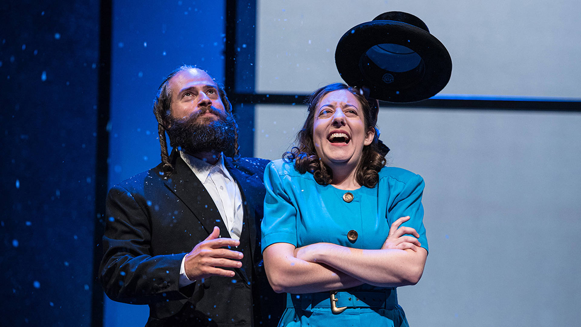 A caucasian man and woman with snow falling around them. The man is dressed in a black jacket and white dress shirt and he has a traditional Hasidic hair and beard. the woman has long brown hair and a blue dress on.