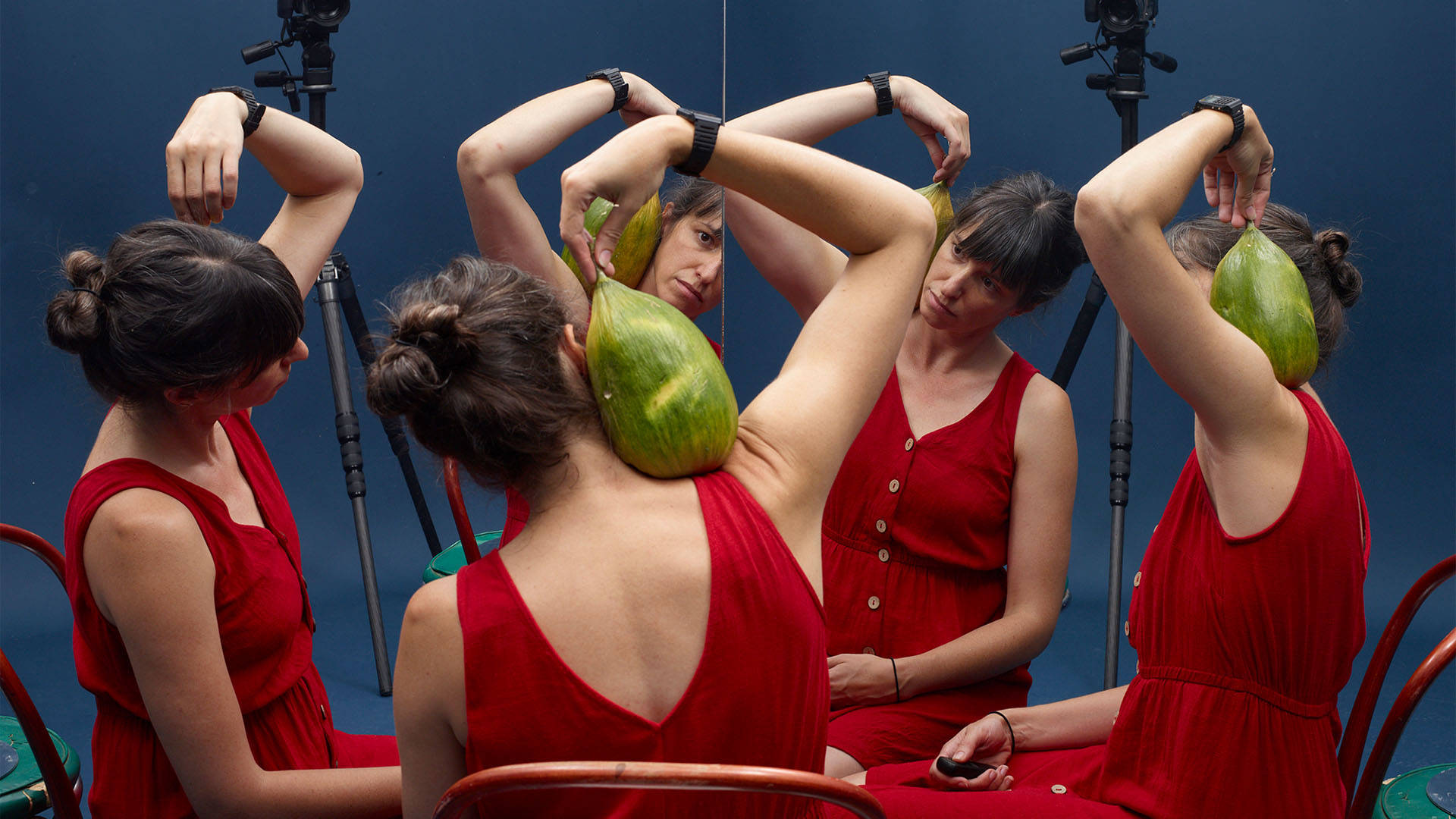 A caucasian woman with dark brown hair in a bun sits in a red button-up dress holding a large green fruit. With her back to the camera, she is facing a multi-direction mirror which splits her image across four reflections.