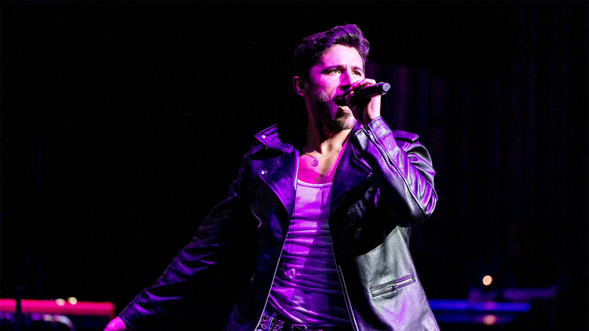 A white man with short brown hair in a leather jacket singing into a microphone.
