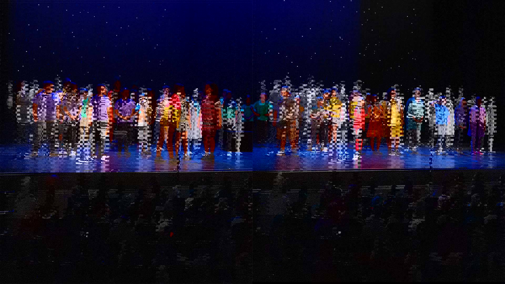 Young students in colorful outfits performing on a dark stage. 