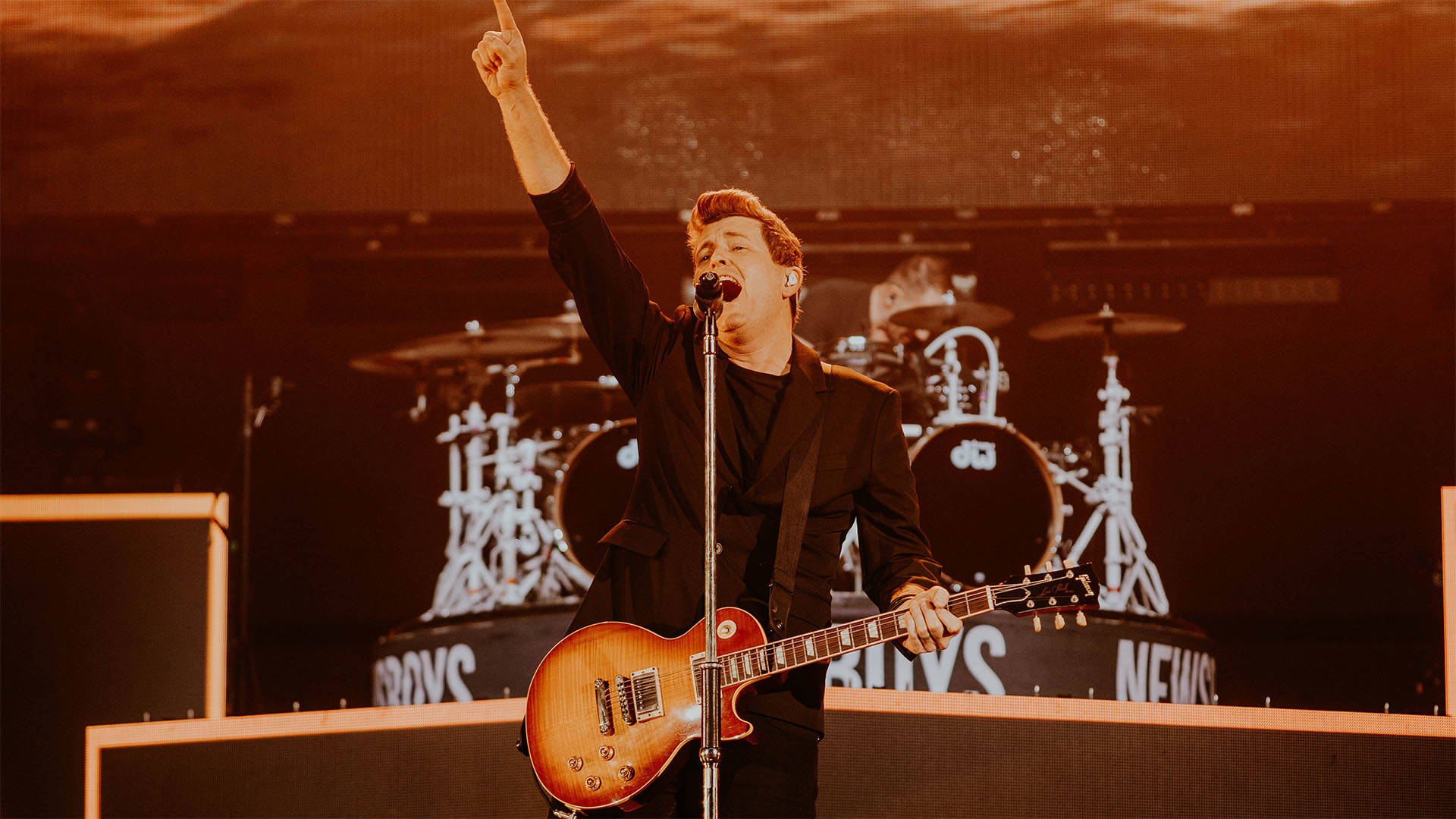 A man playing guitar and singing into a microphone on a stage. He has light complexion and short blond hair and his arm is up in the air.