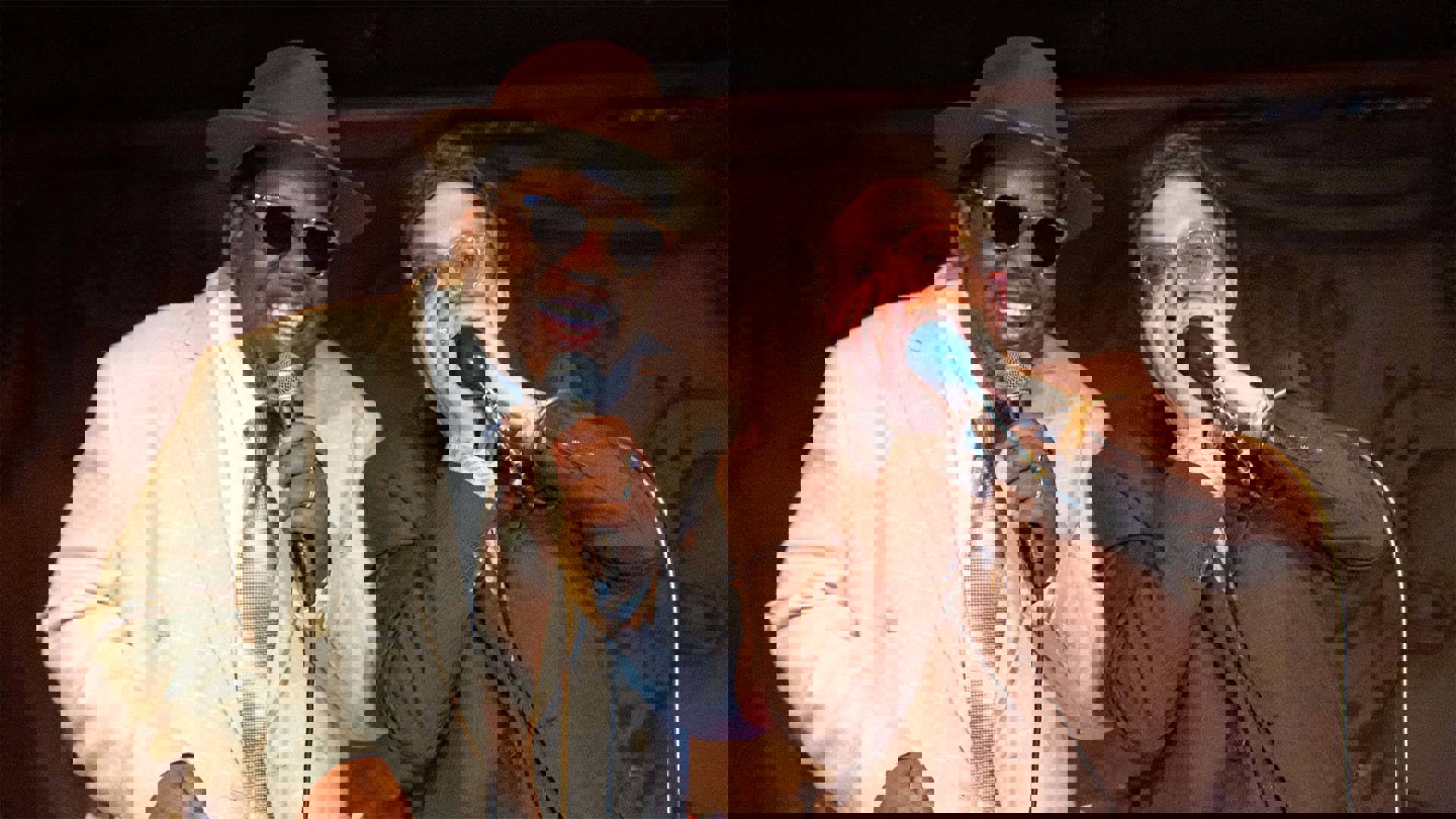 Two men smiling and performing into microphones. The man on the left has dark complexion, a beige suit, brown hat and sunglasses. He is singing. The man on the right has light complexion, a abrown suit, short brown hair and glasses. He is playing a harmonica.