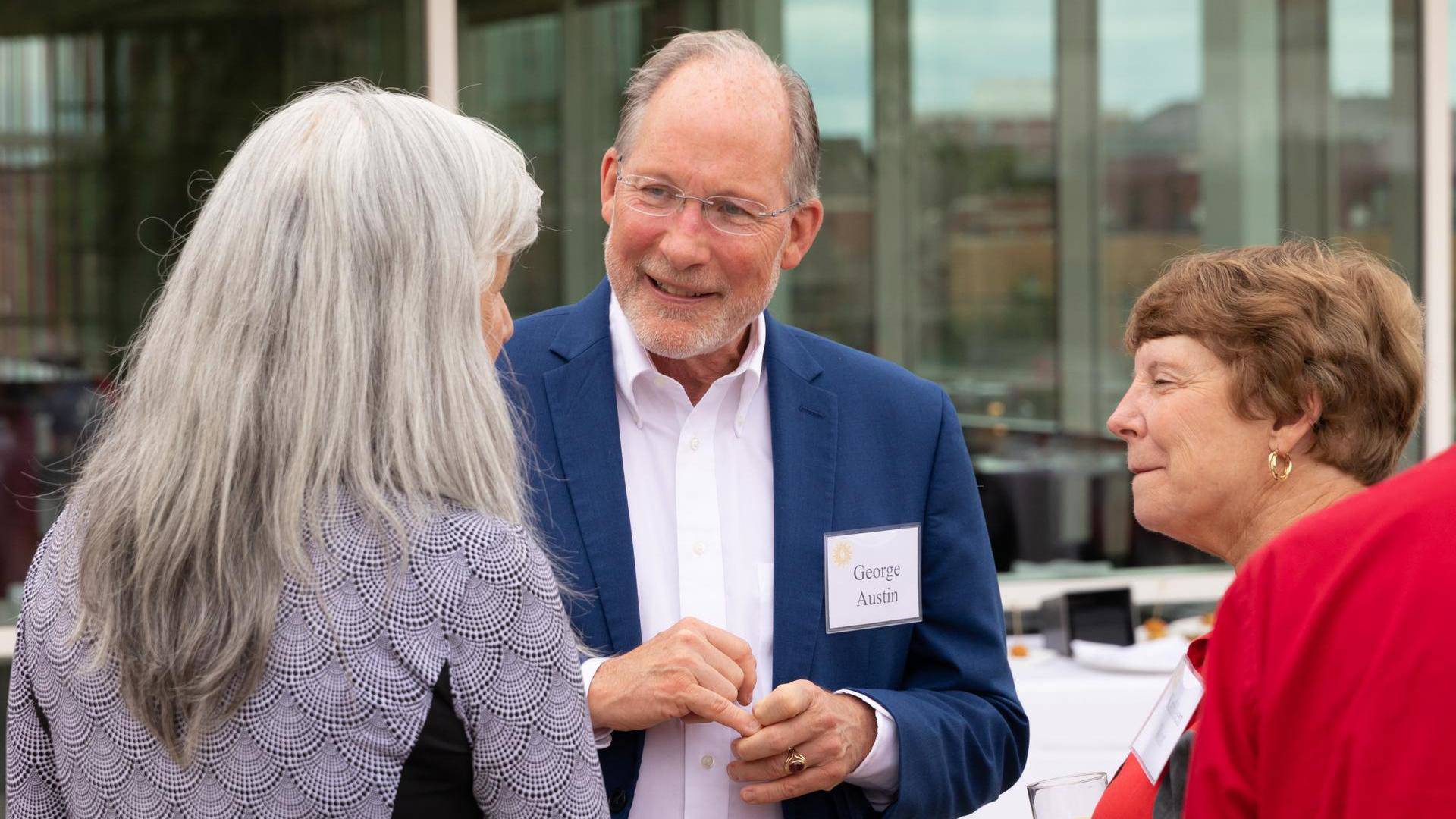 George Austin talk to two women. He is an older caucasian man in a blue suit jacket and white button up. He is wearing thin metal glasses and has grey receding hair with white and gray facial hair.