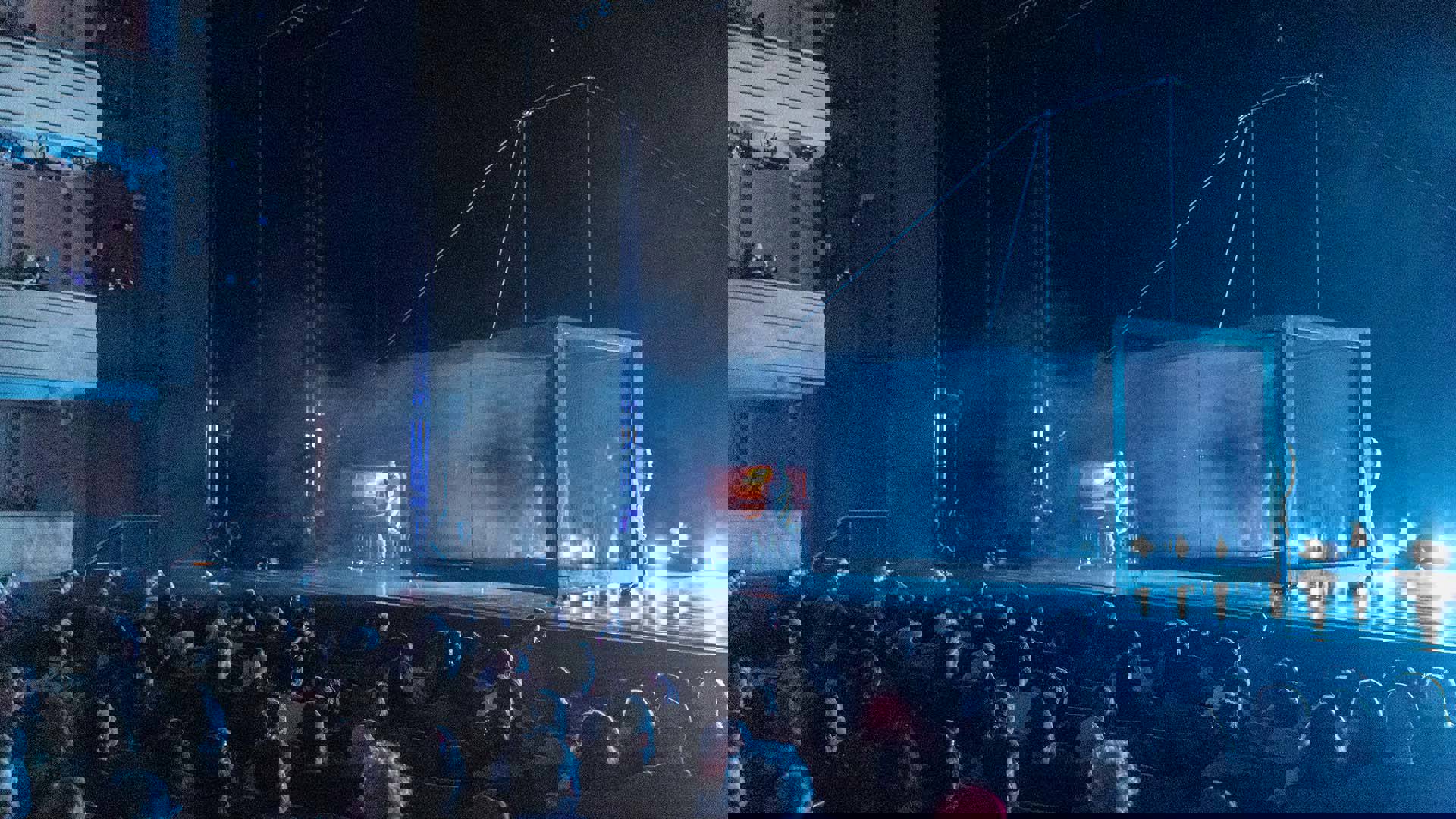 Performers on stage stand inside a foggy glass box while an audience watches.