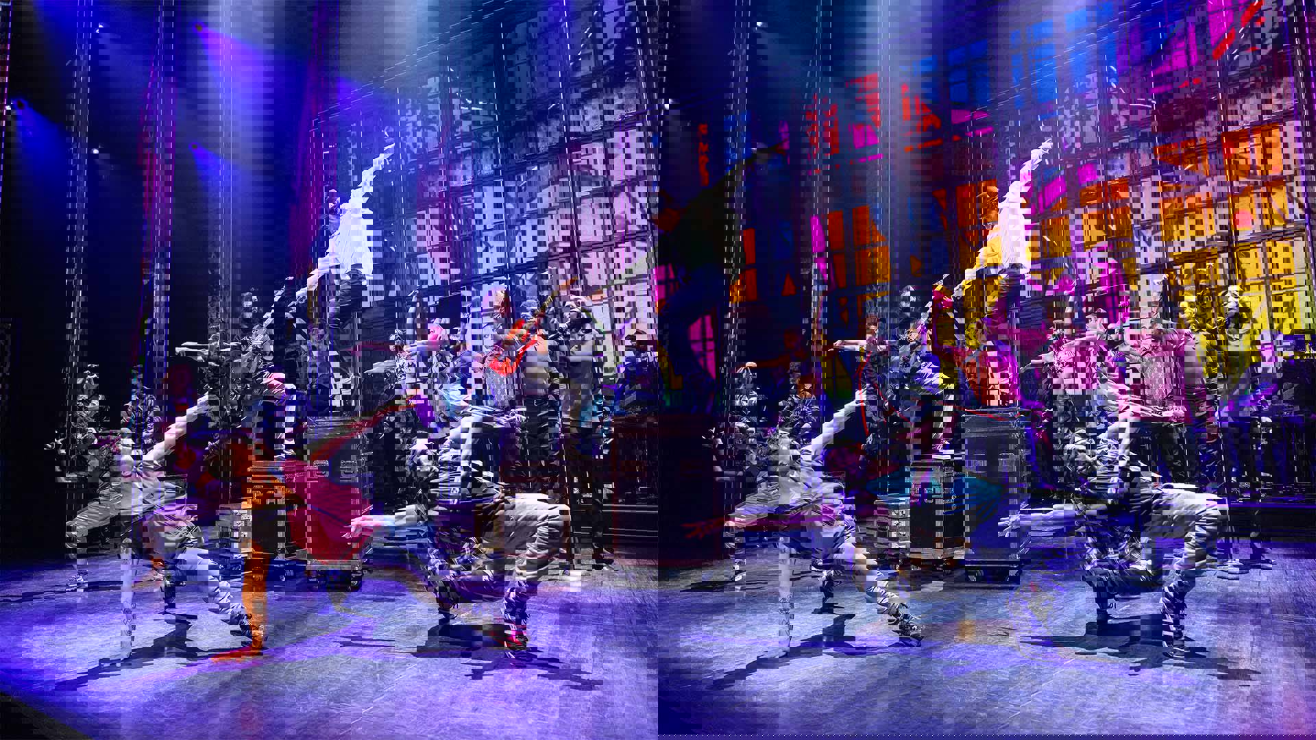 An actor playing Michael Jackson dancing on and equipment box on a stage. He has medium complexion, dark hair, hat, a white shirt, and dark pants. There are other actors dancing around him.