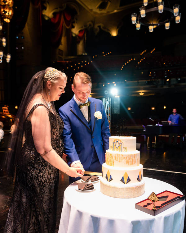 A man in a blue suit and a woman in a beige and black dress with light complexion cutting a wedding cake.