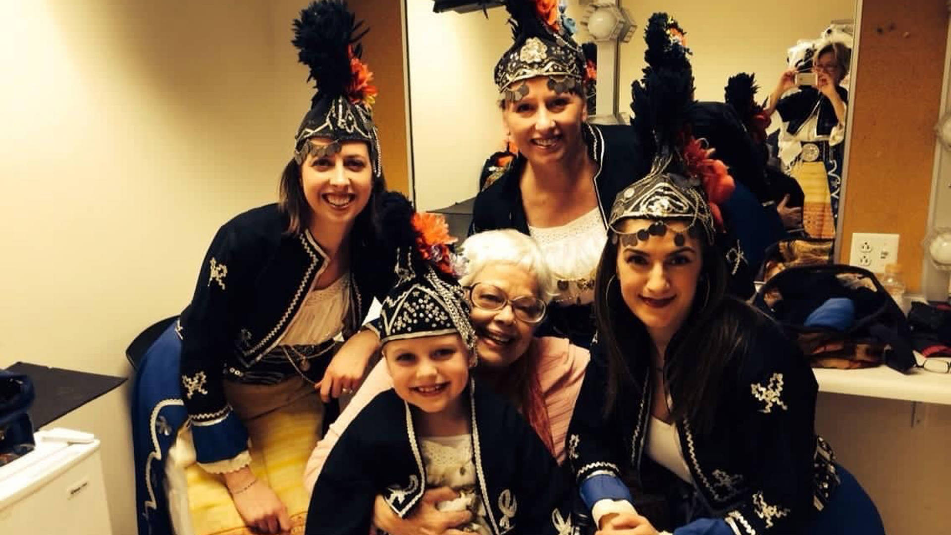 A group of people in traditional Greek dance outfits - black jackets and hats with silver ornaments and red highlights - standing around an older woman with white hair sitting and holding a child on her lap.