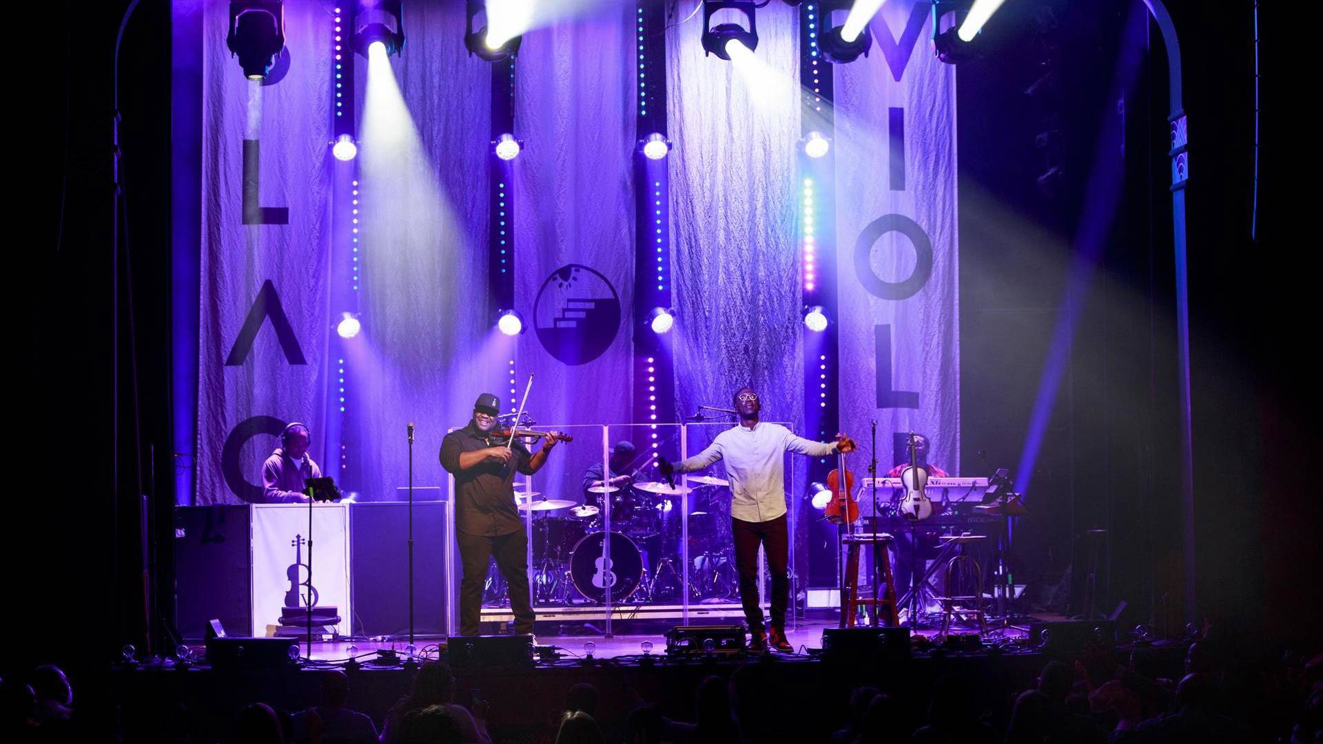 A band performing on stage with two violin players at the front. A keyboard player, DJ and drummer are behind.