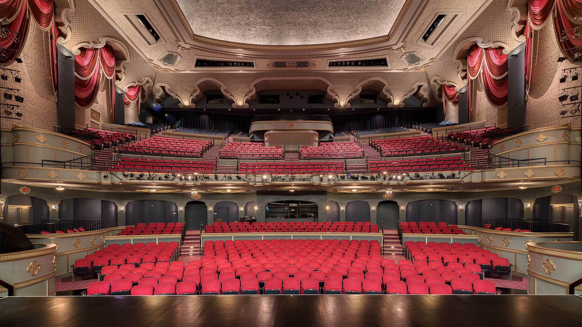 A view from Capitol Theater stage looking out into empty seats of the main floor and balcony. The seats are bright red and the hall is surrounded by red curtains.