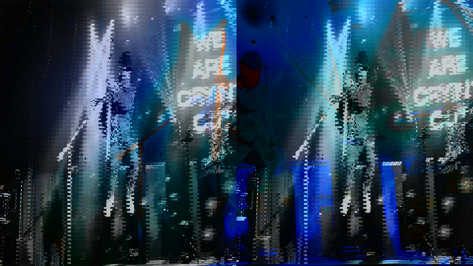 a man in a jean jacket and black hat with a beard and light complexion jumping in the air while he plays guitar.