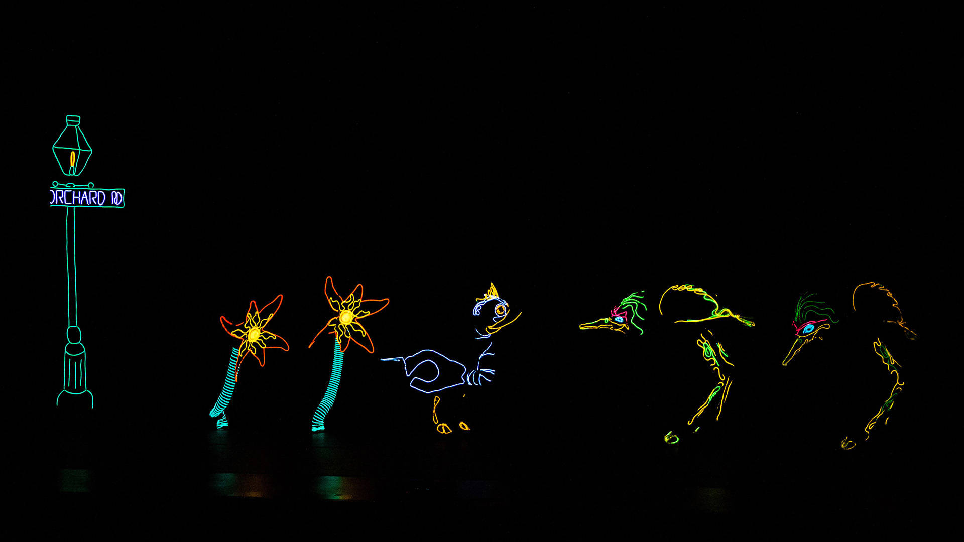 Colorful neon light up puppets and set pieces on a black stage. There are three birds, a lamp post and two flowers.