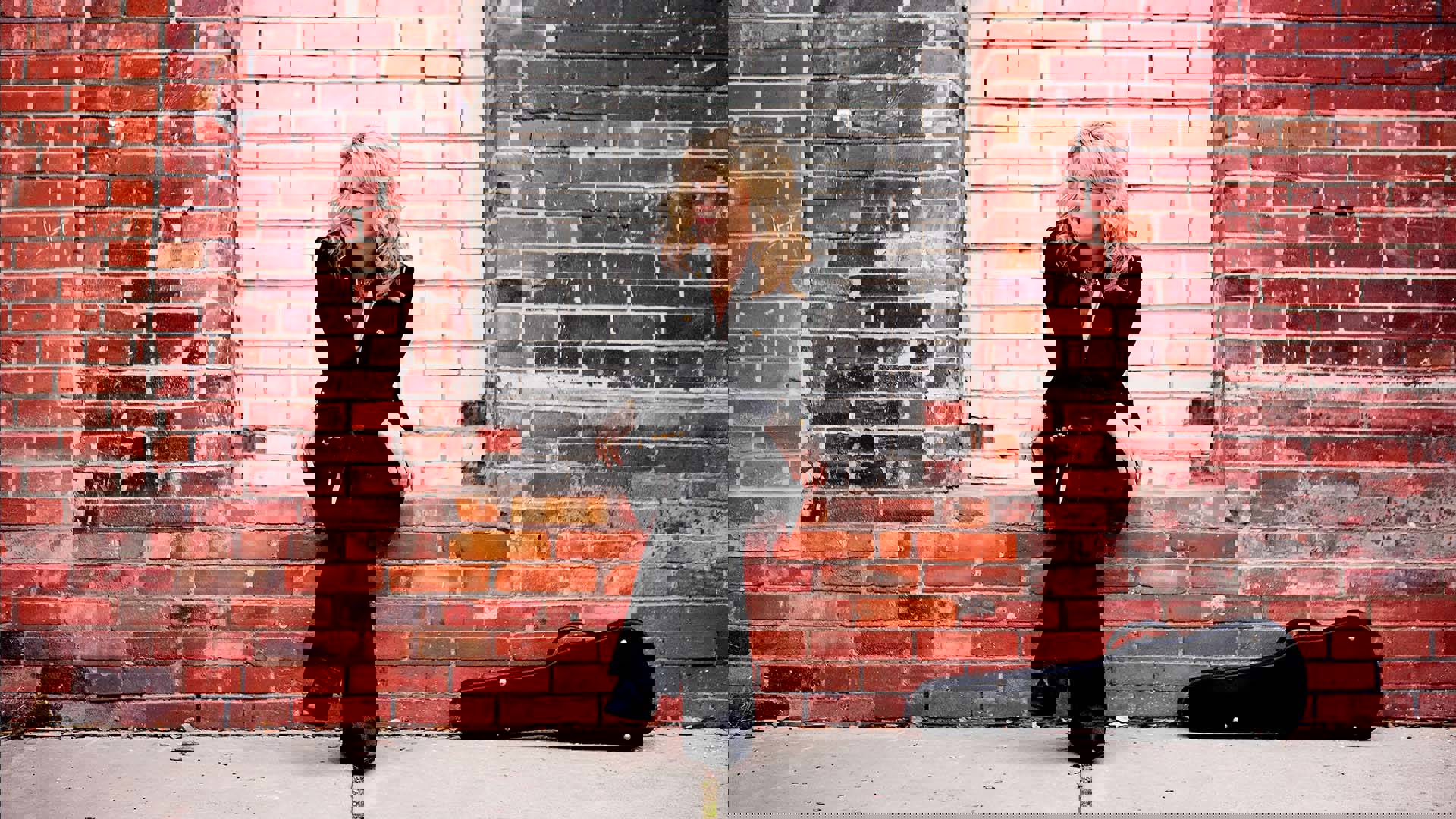 A caucasian woman with blonde hair and a dark outfit sitting in front of a red brick wall. A violin case is on the ground next to her.