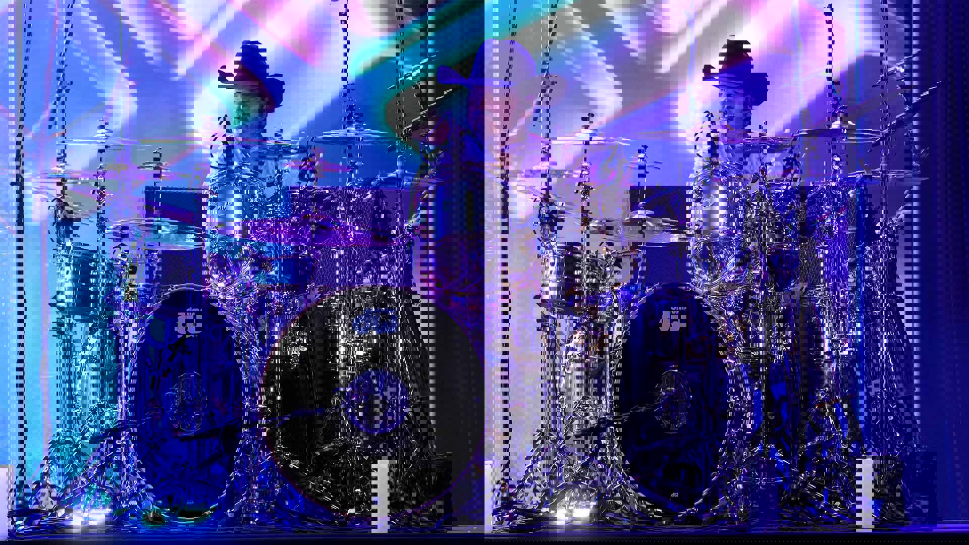 A drummer wearing a black cowboy hat sitting playing a drum kit in front of purple stage lights.