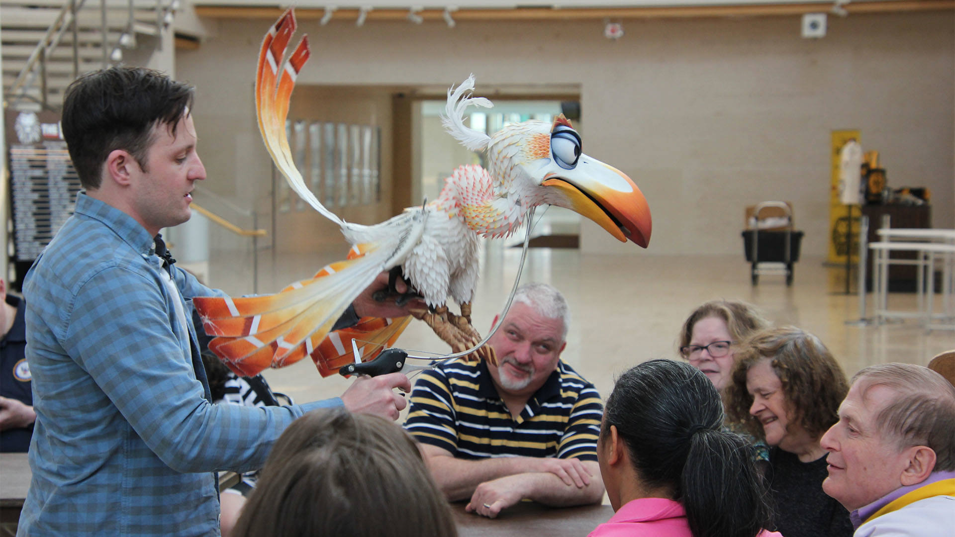 A man in a blue shirt holds a large white and orange bird puppet for a group of people in Overture Hall Lobby.