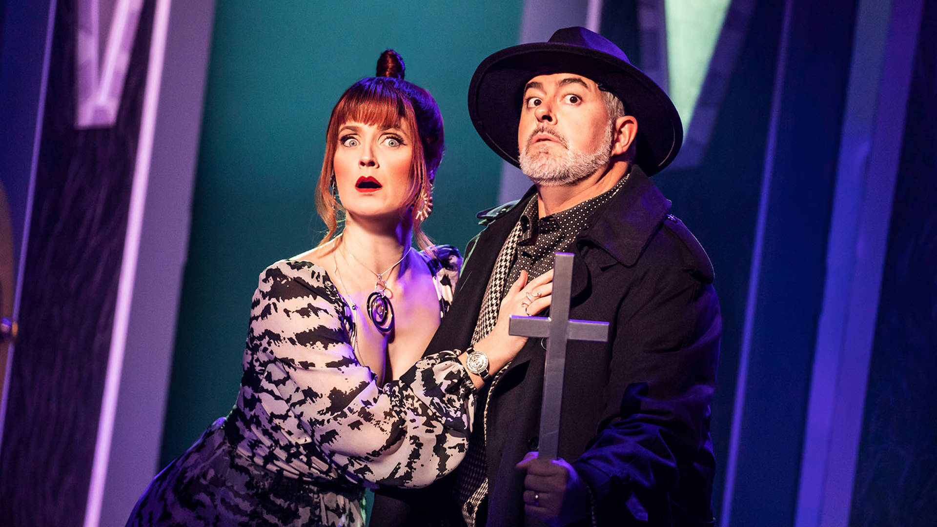 Two people, a woman with red hair in a white & black striped dress and a man in a black trench coat and brimmed hat holding a large cross in his hands, stand in fear.