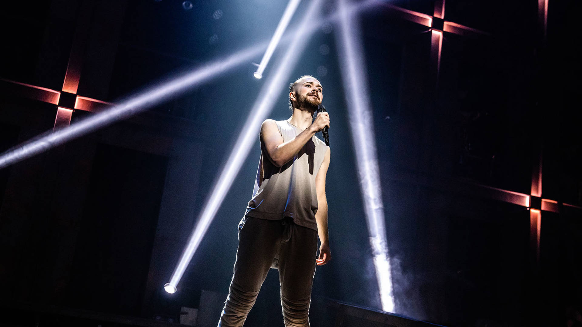 A caucasian man with a beard sings alone on stage.