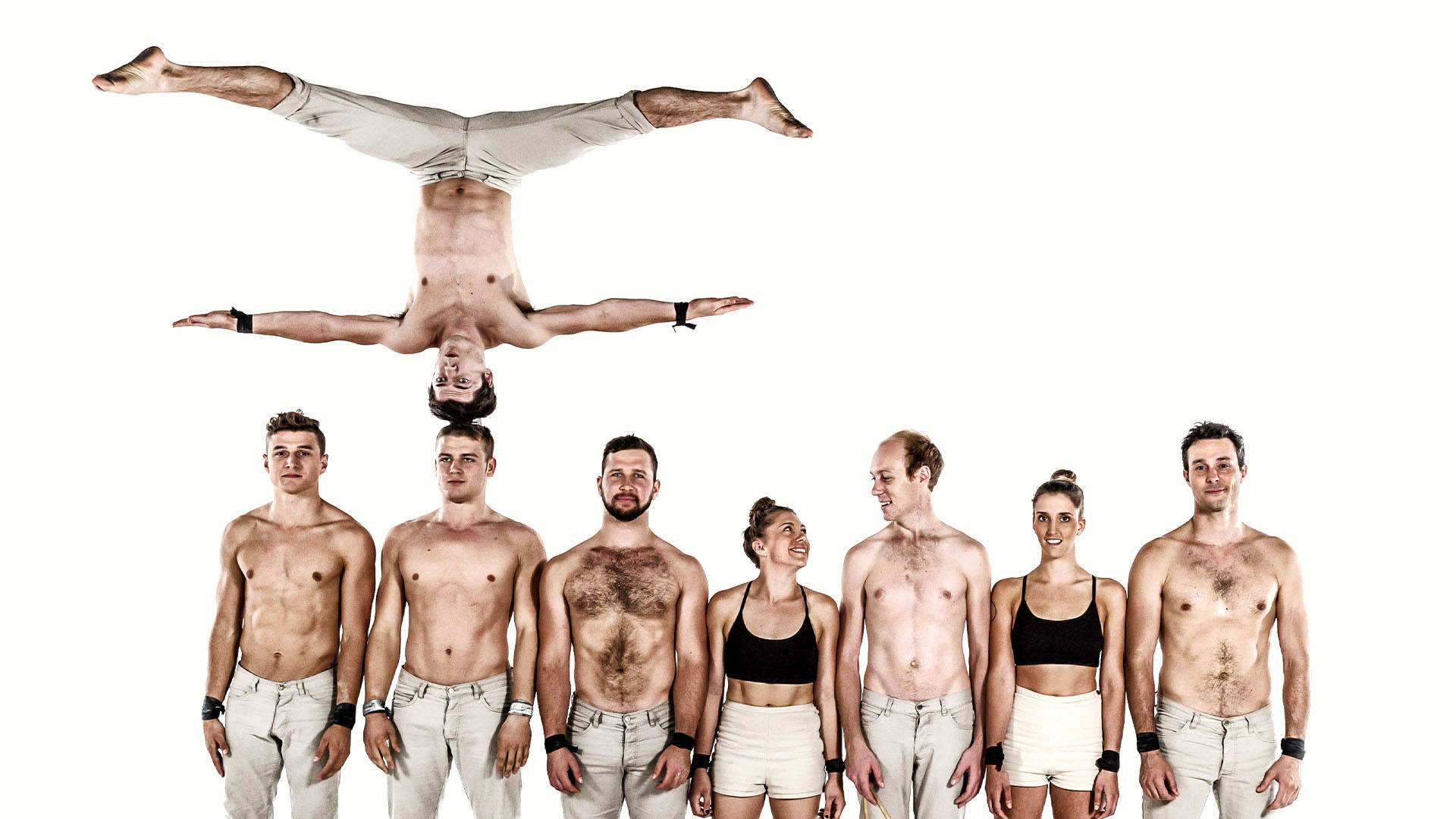 A group of performers stand in a single file line, shoulder to shoulder. The women are in tanktops and the men are shirtless. A single man is balancing upside down on another man's head.